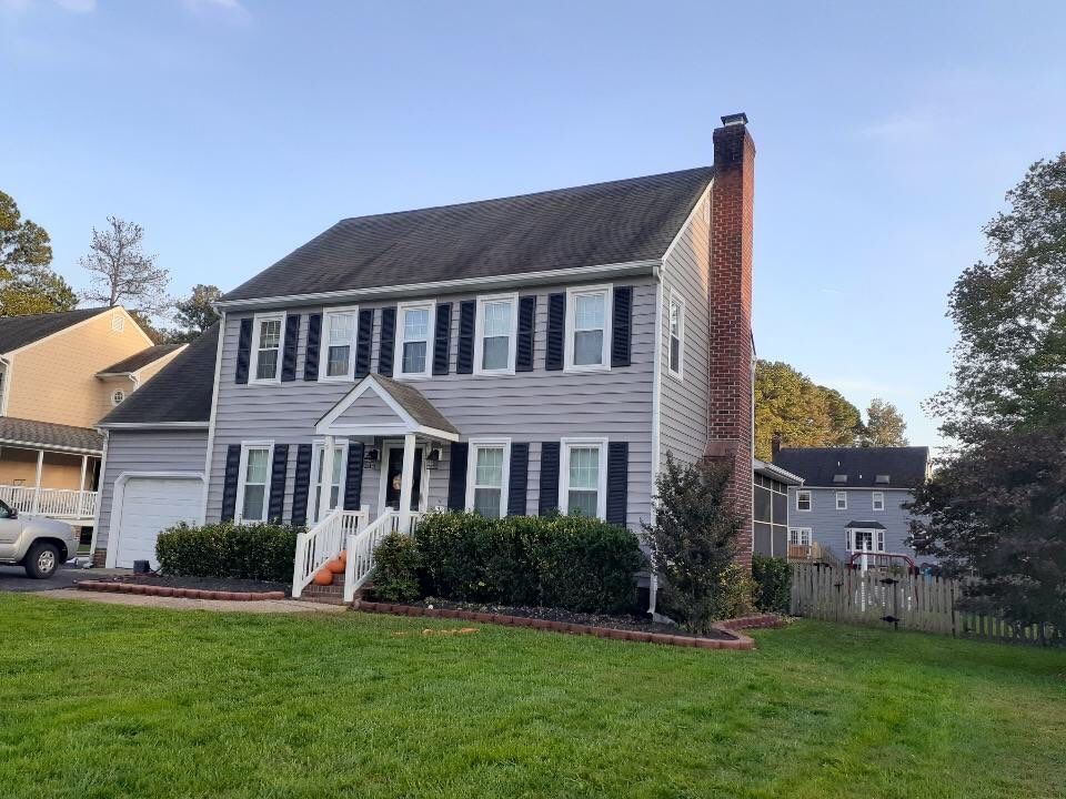 Two-story gray house with black shutters, a red-brick chimney, and a small porch; green lawn in front.