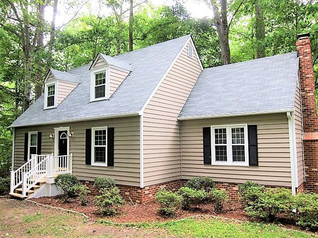Beige house with gray roof, black shutters, and brick chimney surrounded by trees.