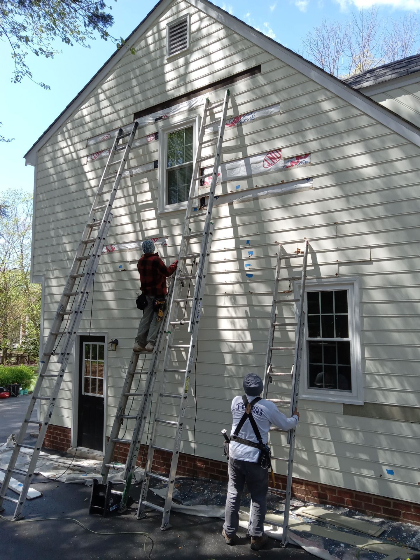 Three workers on ladders siding a light-colored house. One person is actively working near a window.