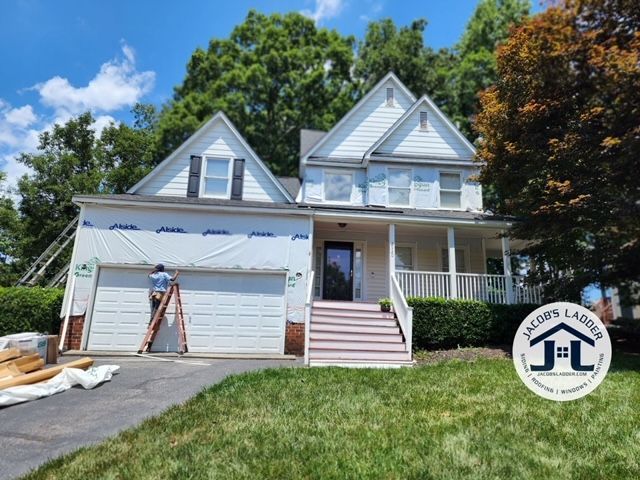 House undergoing siding work; worker on ladder, garage door, porch, blue sky.