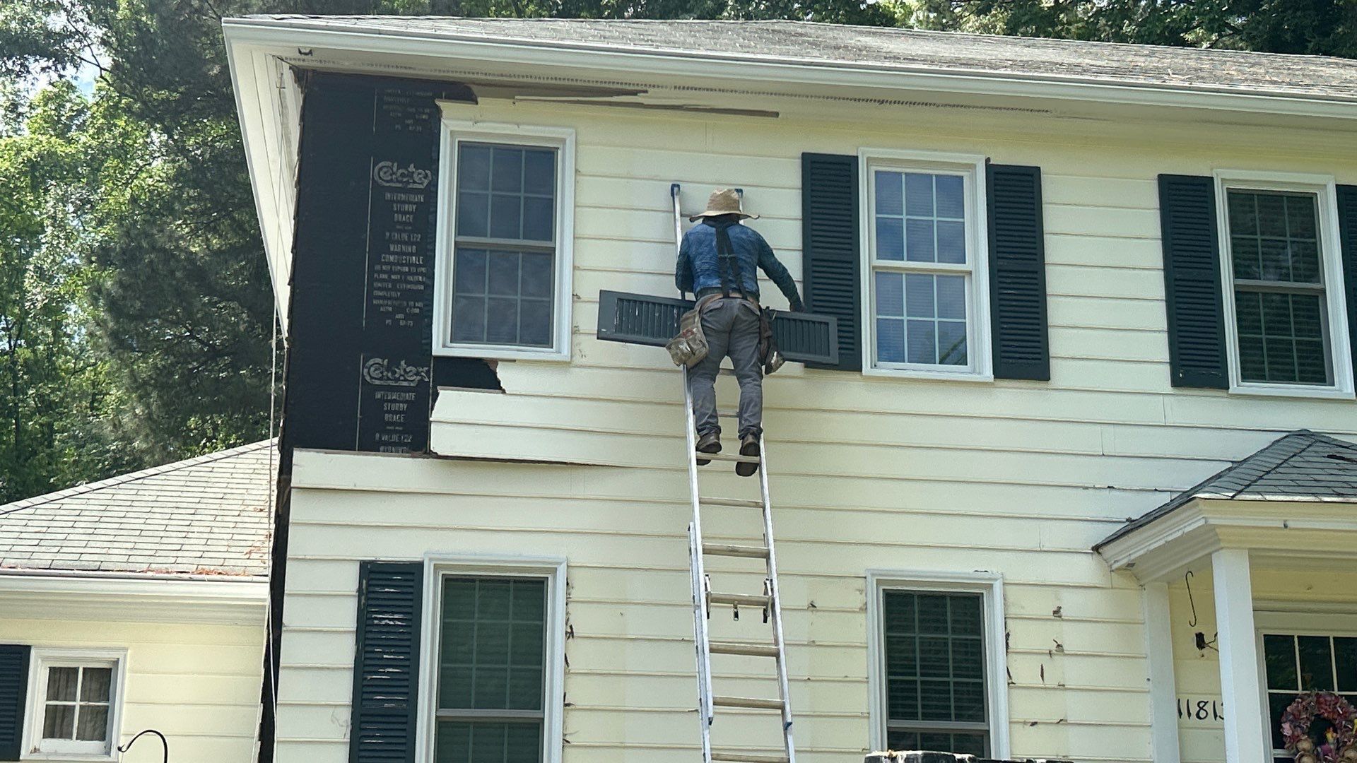 Person on a ladder removing siding from a two-story house with black shutters; exposed black siding.
