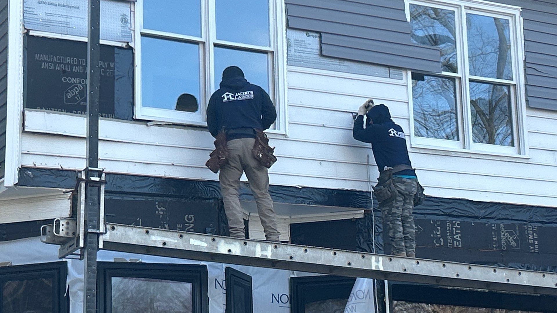 Two workers siding a house, standing on a ladder. One inside a window frame, other using a drill.