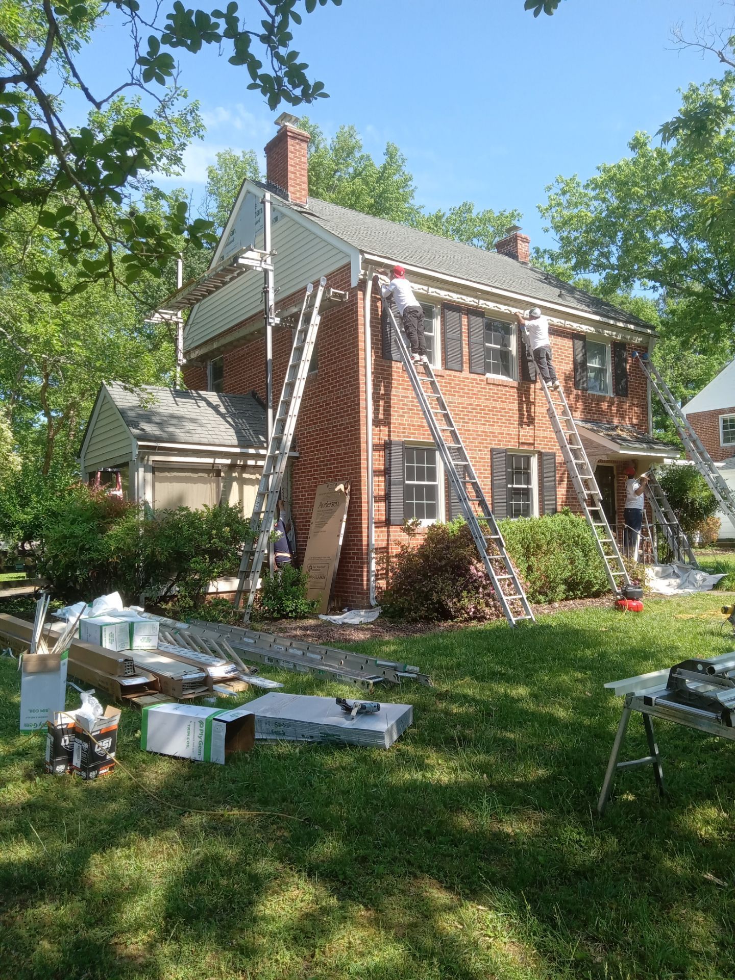 Workers on ladders installing siding on a two-story brick house with a green lawn. Blue sky, tools visible.