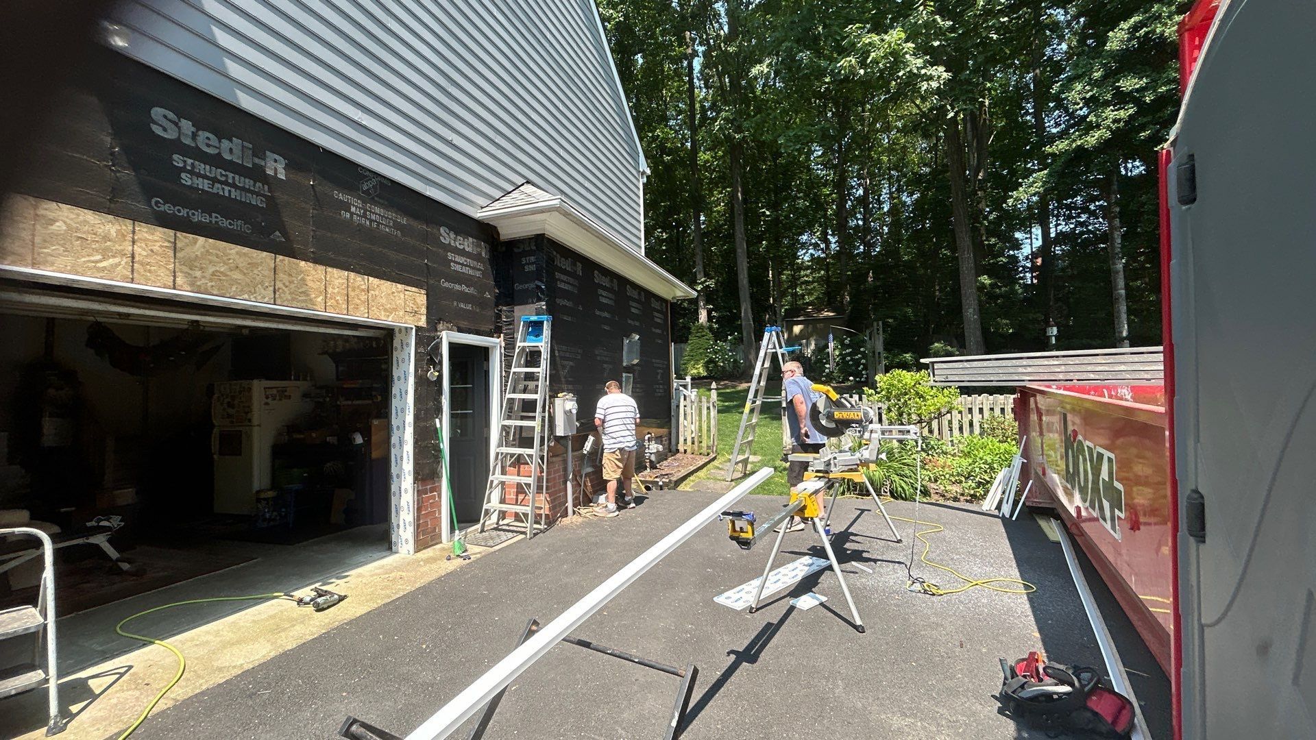 Construction workers outside a garage, working on the building's exterior. Siding and tools are visible.