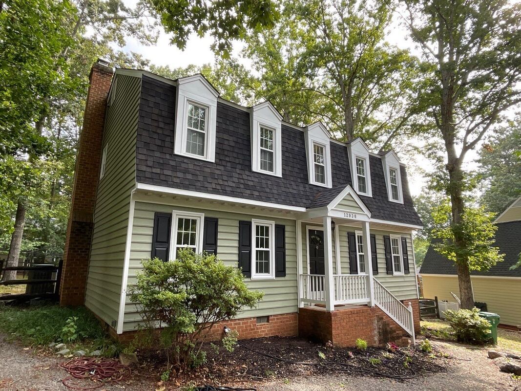 Green house with black shutters, dark roof, dormer windows, and brick chimney.