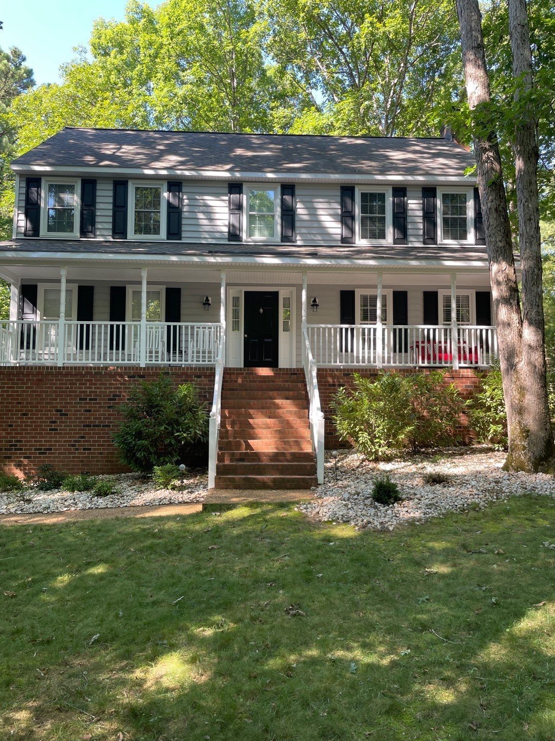 Two-story house with gray siding, black shutters, white porch with railing, brick base, and front steps.