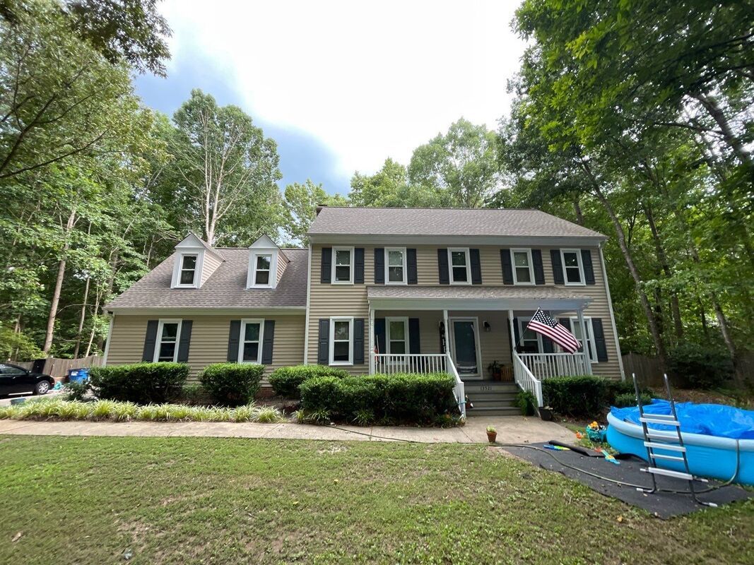 Two-story house with tan siding, black shutters, and an American flag. Lawn and trees surround.