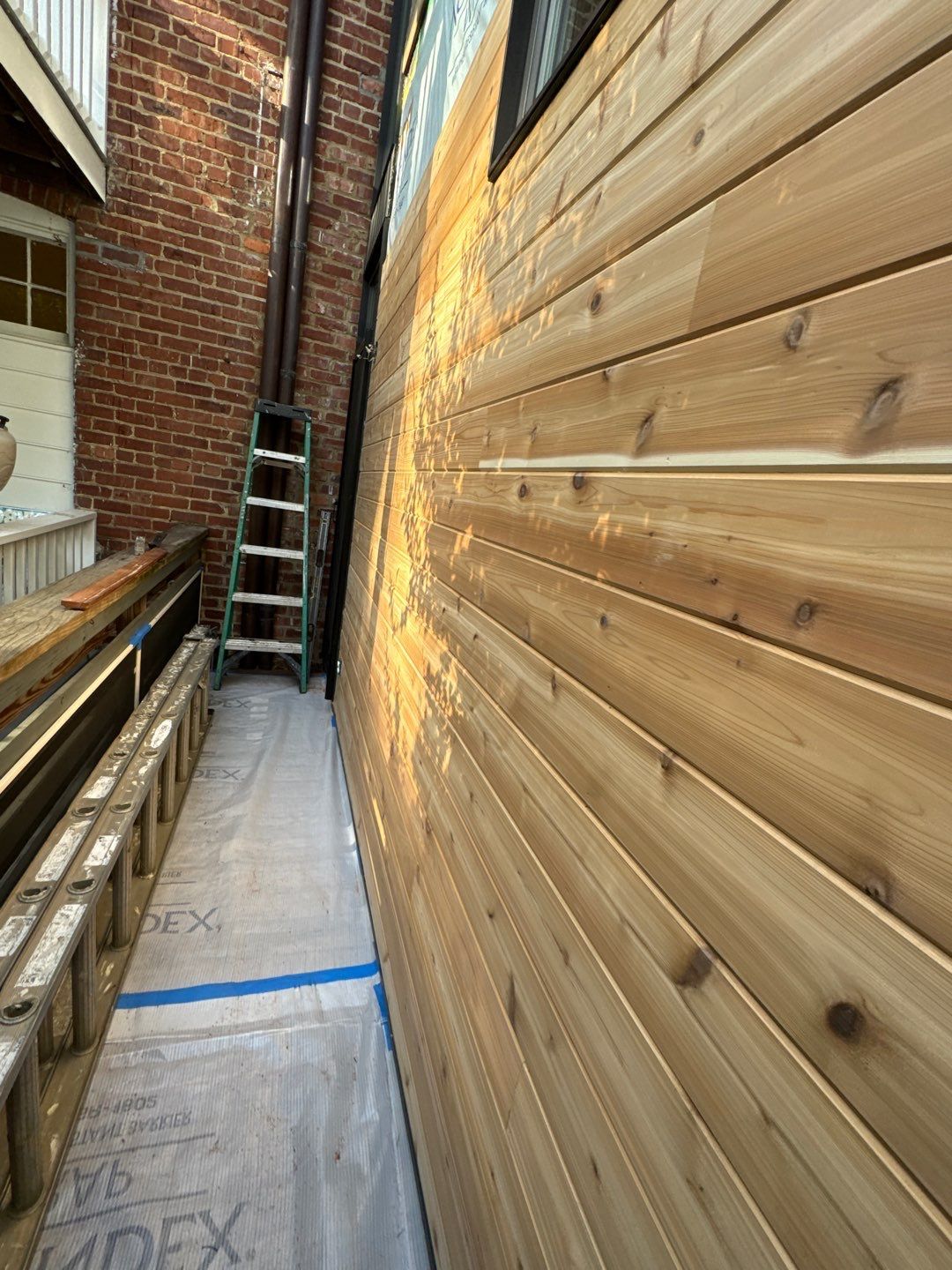 Cedar siding being installed on a building exterior next to a brick wall. A ladder and lumber are nearby.