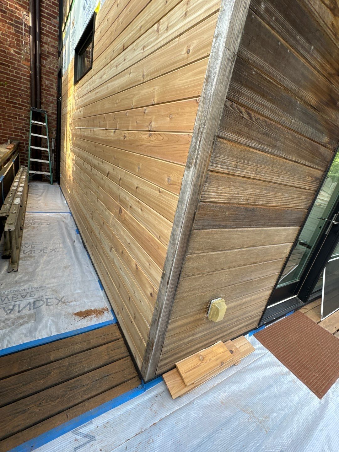 Wooden-sided triangular building under construction. Brown siding, white protective covering, and a ladder are visible.