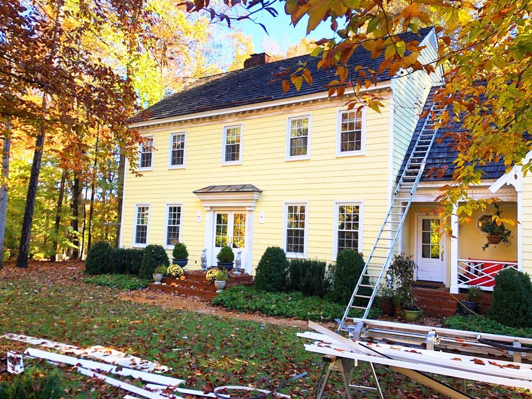 Yellow house with a dark roof and white trim, surrounded by fall foliage. A ladder leans against the side.