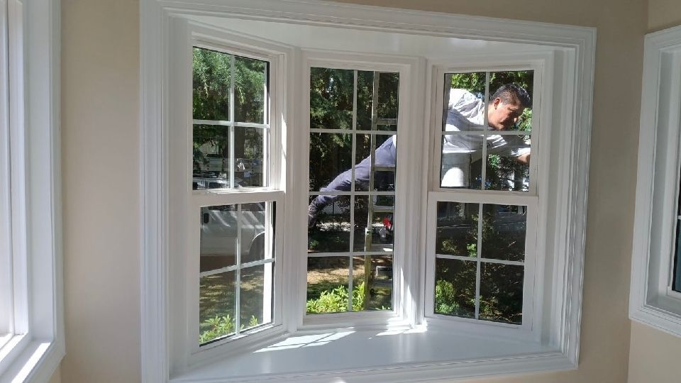White bay window with grids; person outside cleaning, reflections of trees and vehicles.