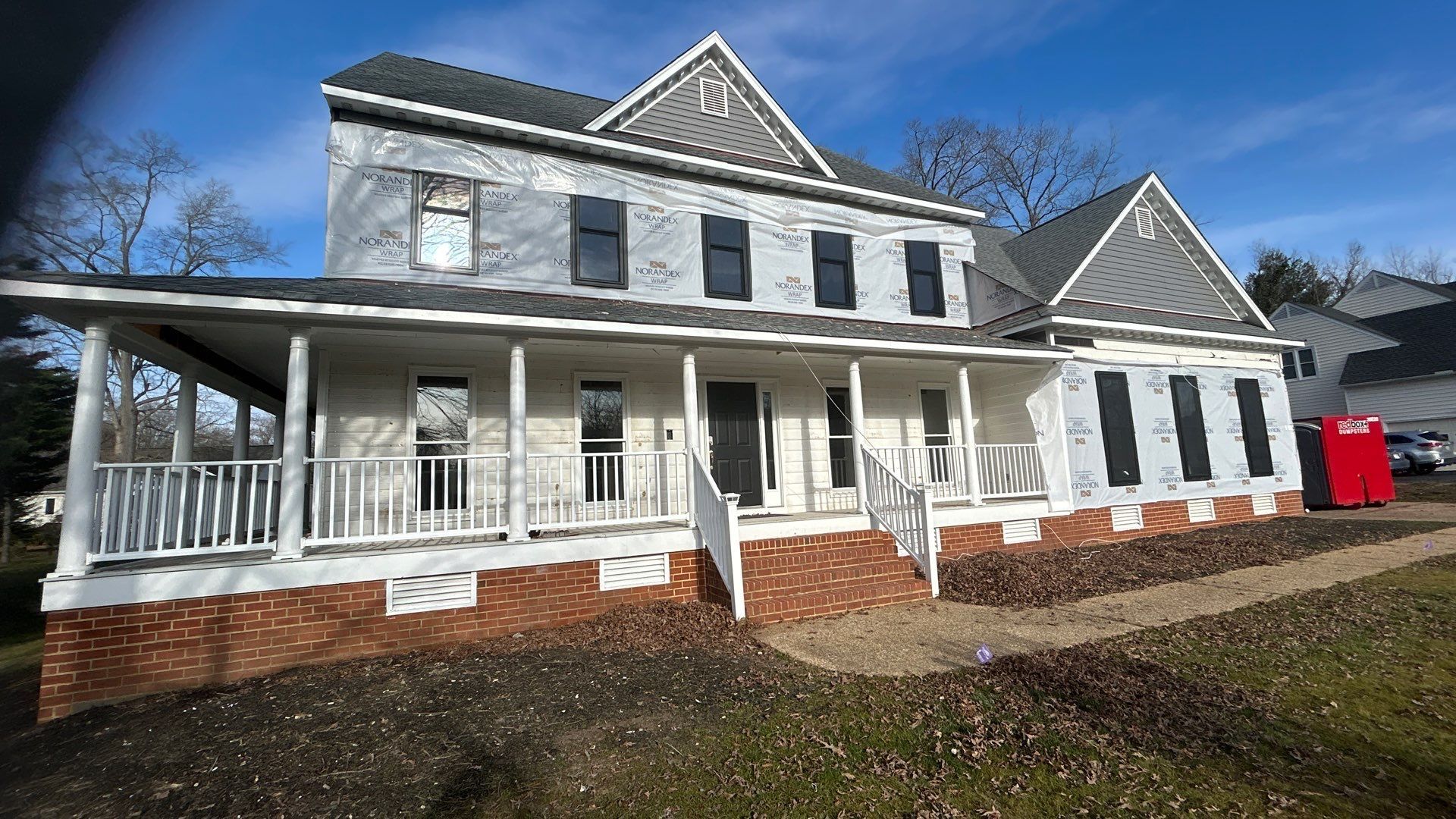Two-story house with white trim, gray siding, covered porch, and brick foundation, under a blue sky.