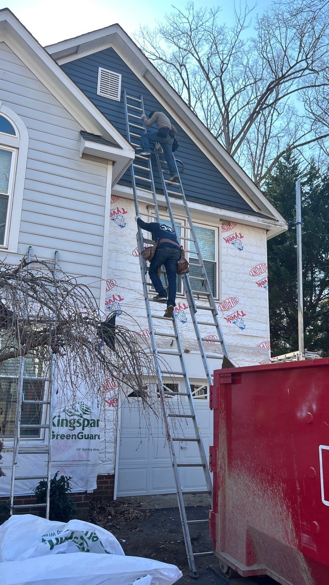 Two workers on ladders siding a house, white siding and blue gable. Red dumpster nearby.