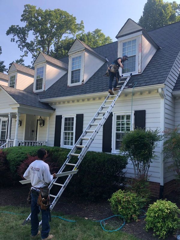 Two workers on a tall ladder, one near an upper window, on a white house with black shutters, roof work.