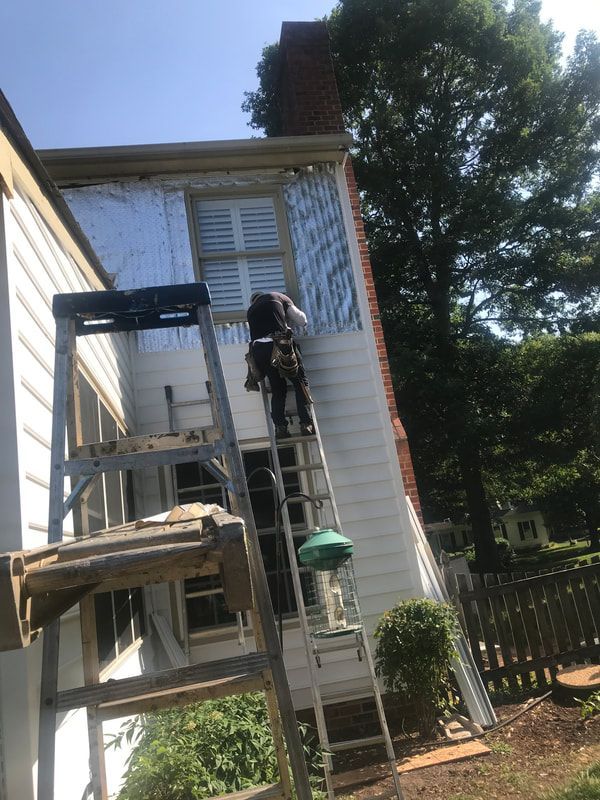 Person on ladder working on the side of a white house with insulation. Blue sky, brick chimney.