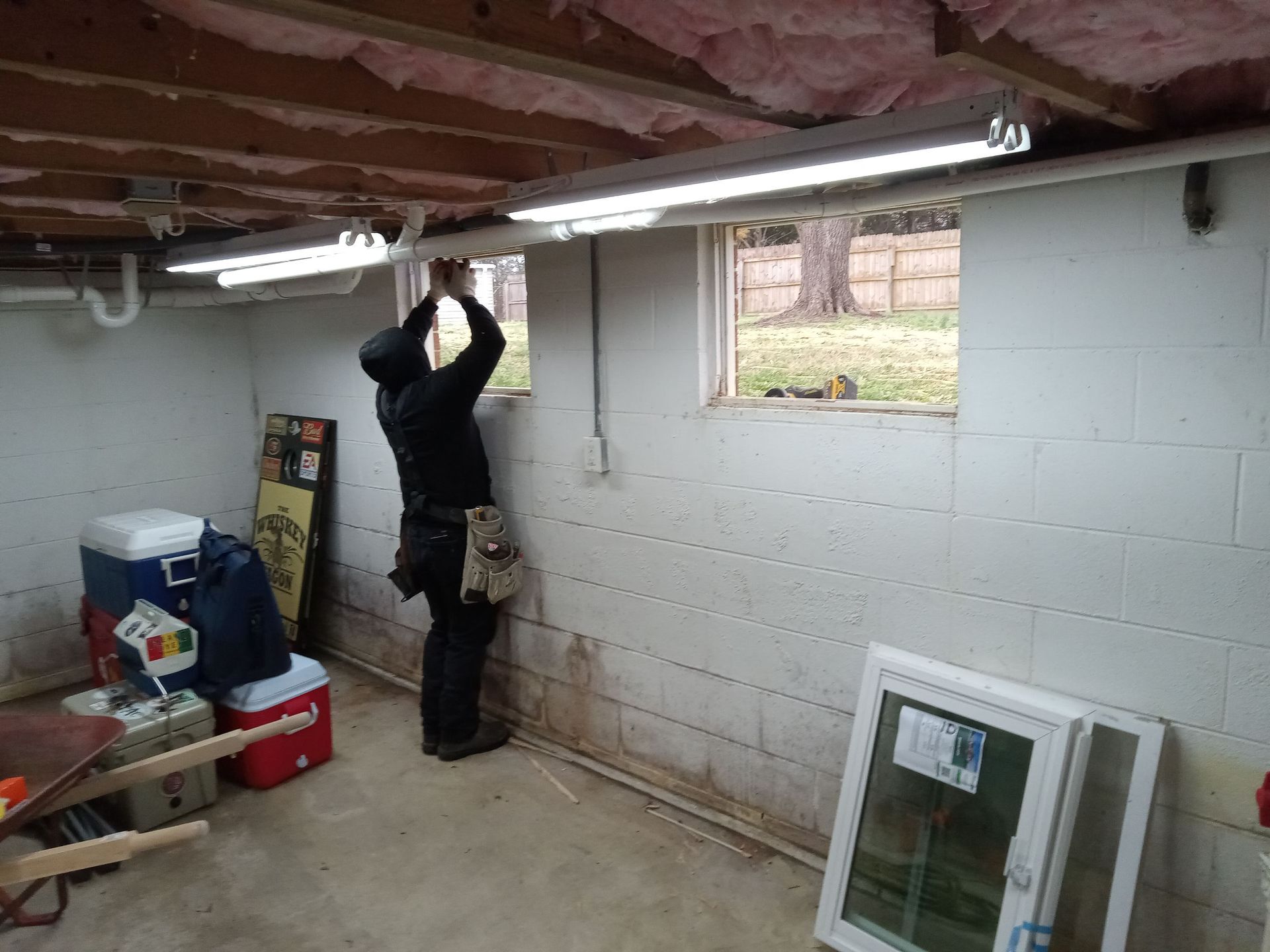 A person installing fluorescent lights in a basement with a window. White walls, concrete floor.