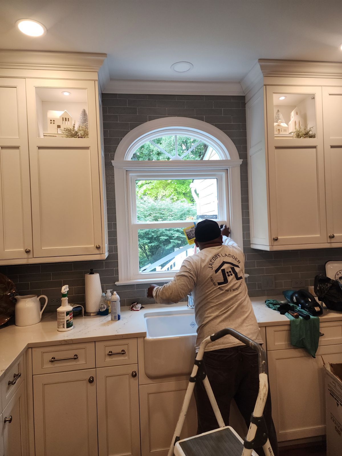 Man on ladder installing window in kitchen with white cabinets.