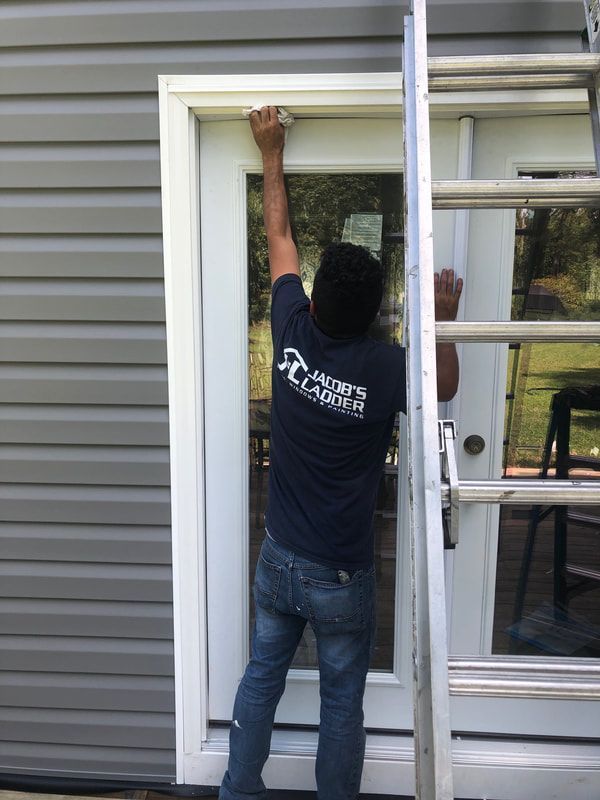 Person on ladder installing something above a white door. Gray siding, blue shirt, jeans.
