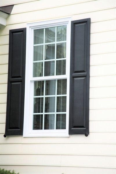 Window with black shutters on a cream-colored house, white trim, blinds visible through the glass.