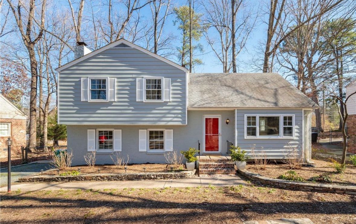 Two-story light blue house with white shutters and a red door. Trees surround the building.