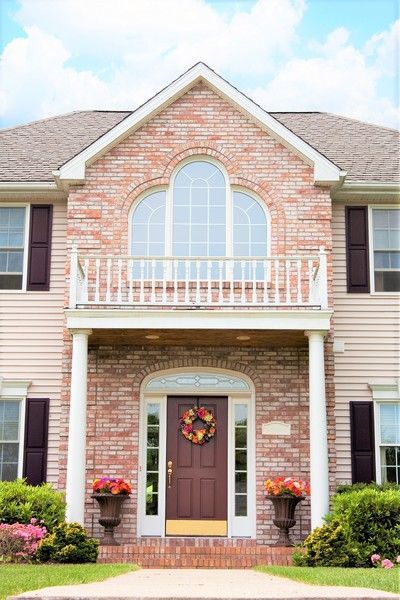 Two-story house with brick facade, balcony, burgundy door, flower pots, and a wreath.