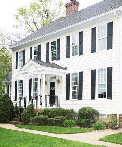 Two-story white house with black shutters, black railing, and a small front yard with green bushes and grass.