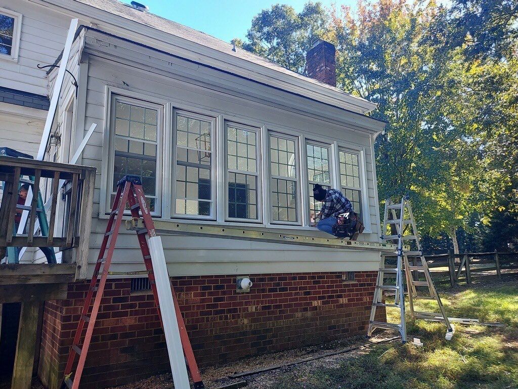 Person working on the exterior of a house with windows; two ladders and building materials visible.