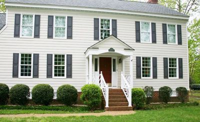 Two-story beige house with black shutters, white trim, and a red front door. Brick steps lead to the porch.