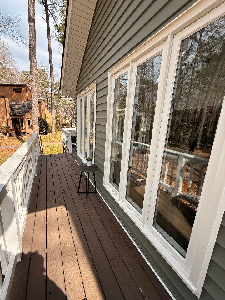 Exterior view of a house with a deck. The house has green siding and large windows, and the deck has brown planks and white railings.