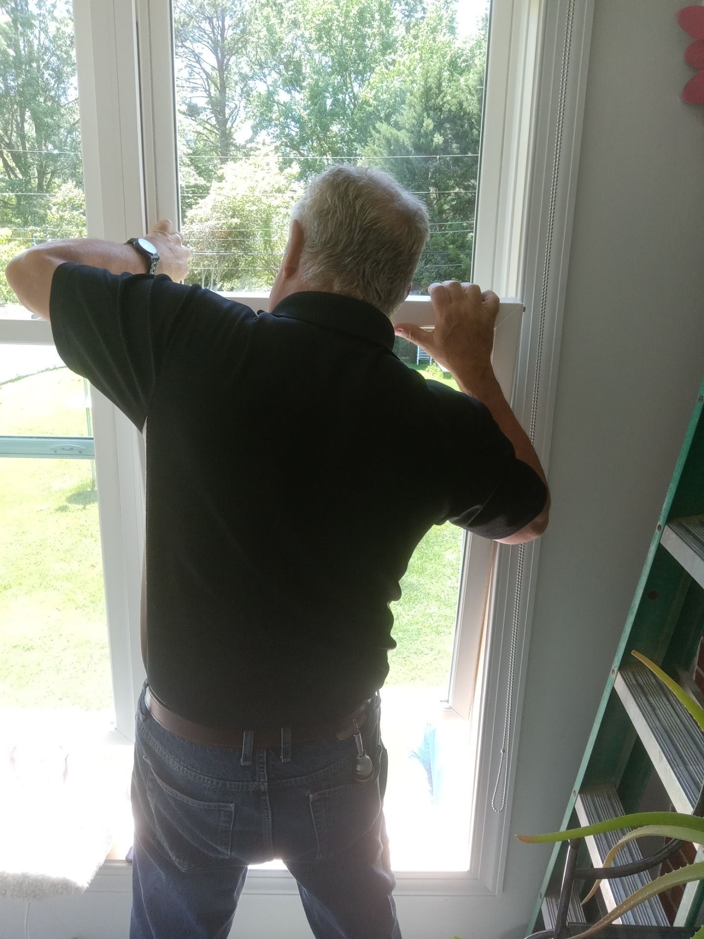 Man working on a window from the inside. He wears a black shirt and jeans. Bright outdoor light shines through.