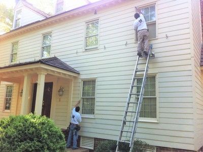 Two people painting the exterior of a two-story beige house. One is on a ladder, the other is on the ground.