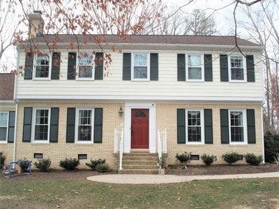 Two-story beige house with red door, black shutters, and tan brick facade.
