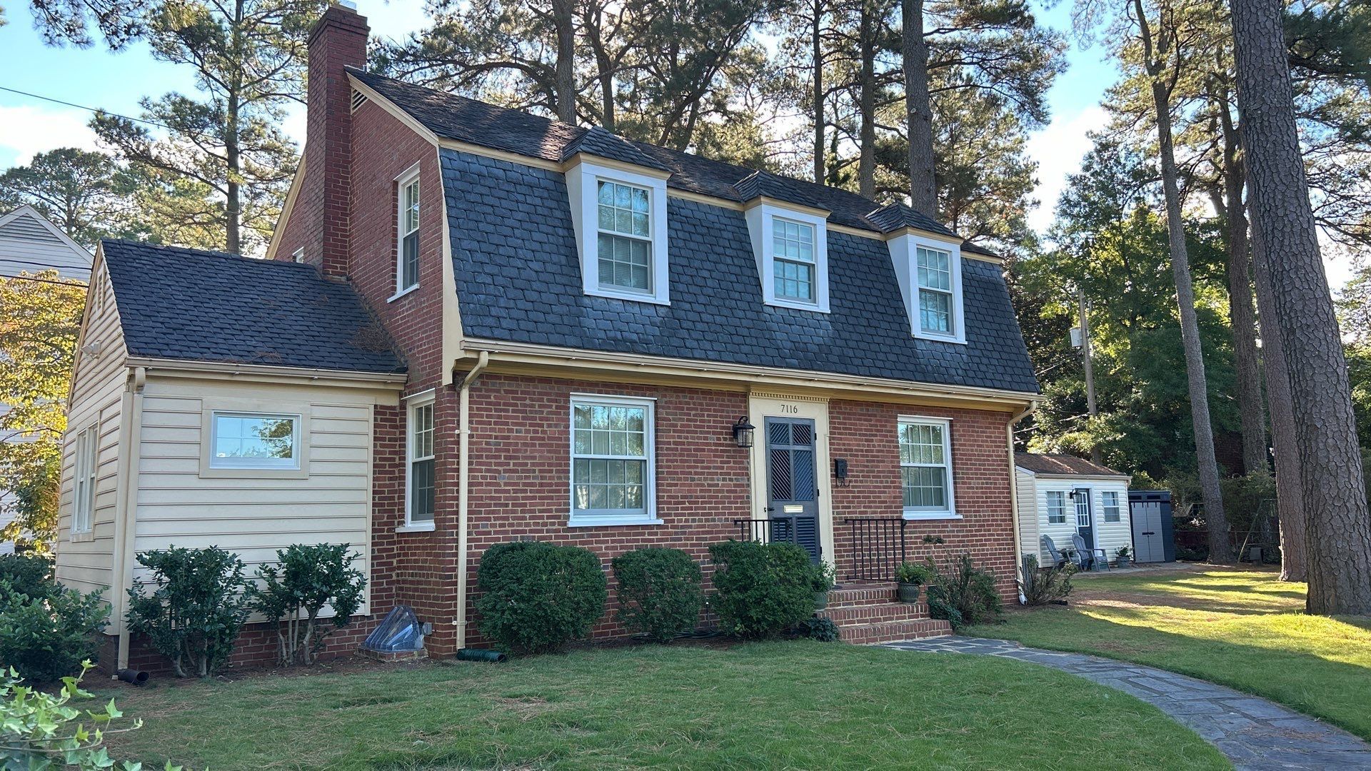 Brick house with dark roof and dormer windows, surrounded by trees and a green lawn.