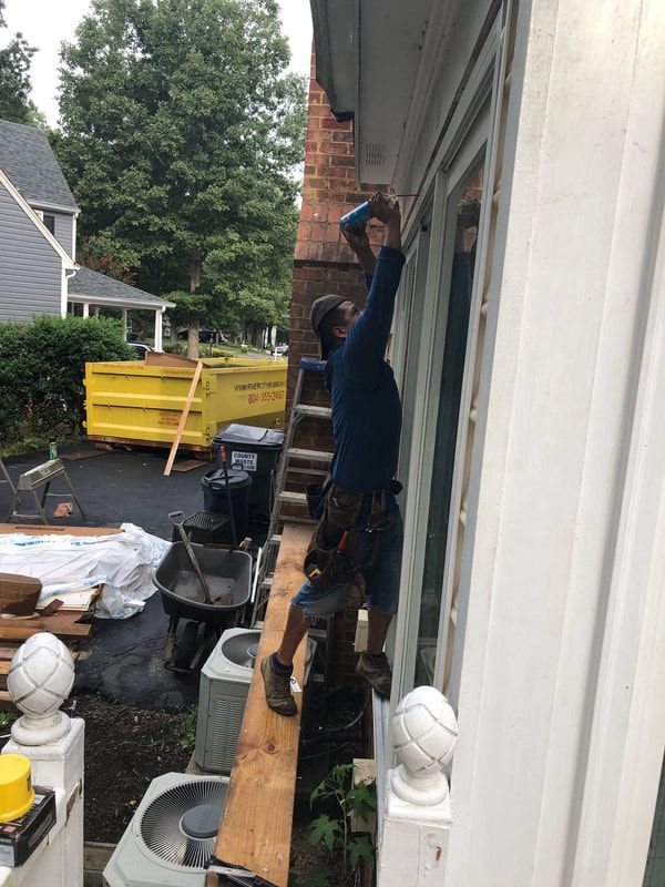Man on ladder installing window trim on a house with a brick wall.  Outdoor setting; yellow dumpster.