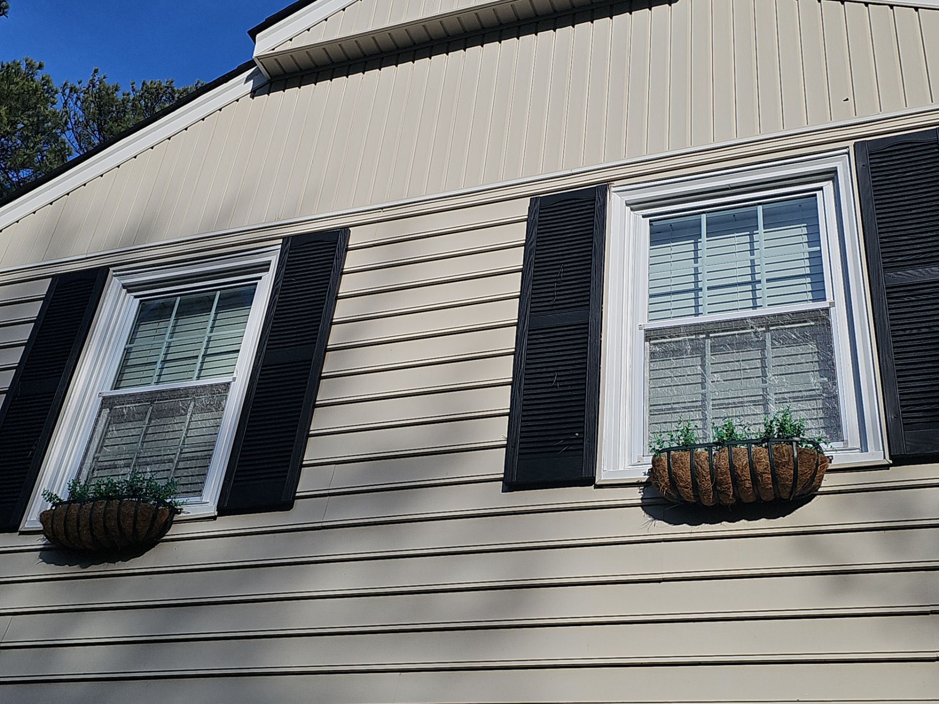 Two windows with black shutters, hanging flower baskets, on a beige-sided house under a blue sky.