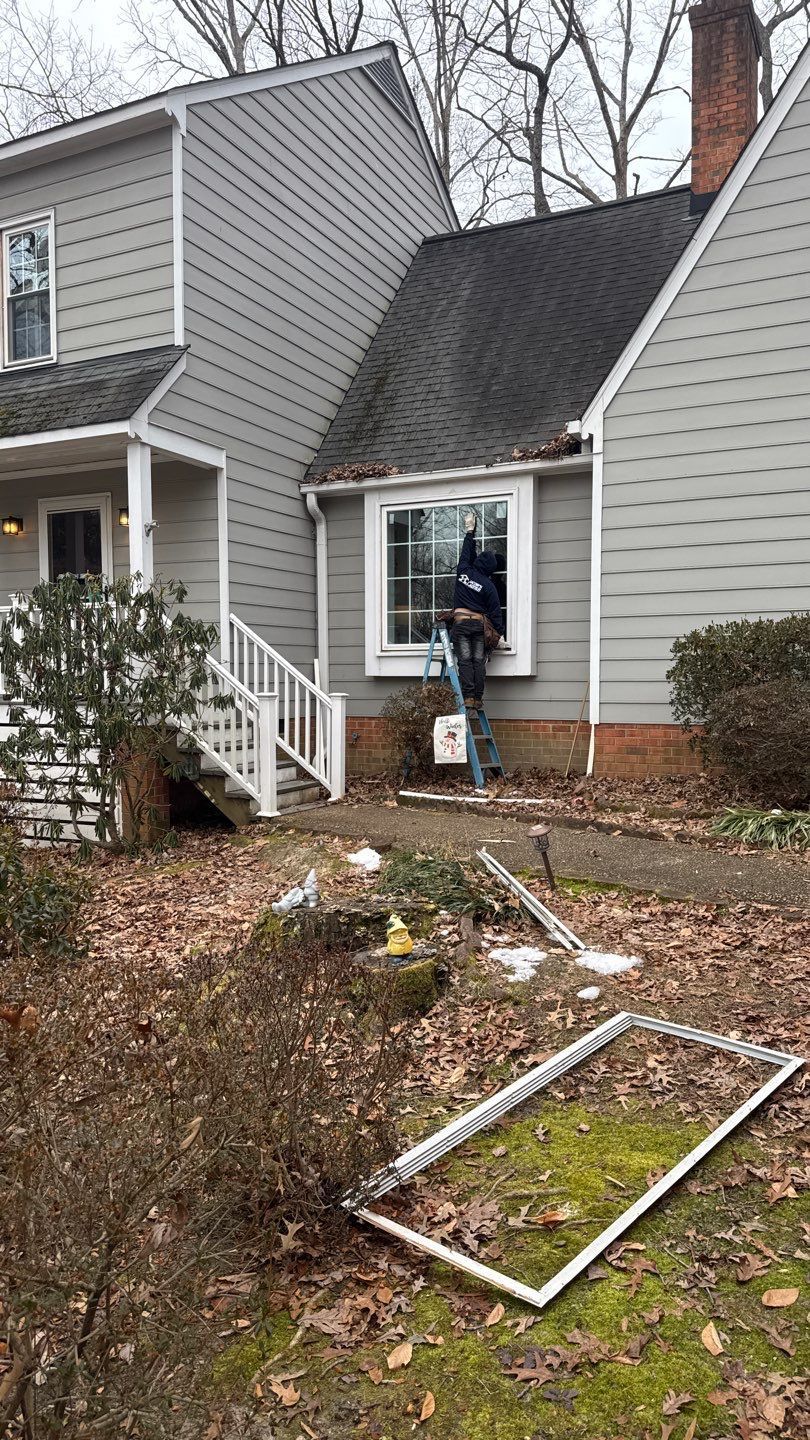 A person on a ladder working on a window of a gray house; debris on ground.
