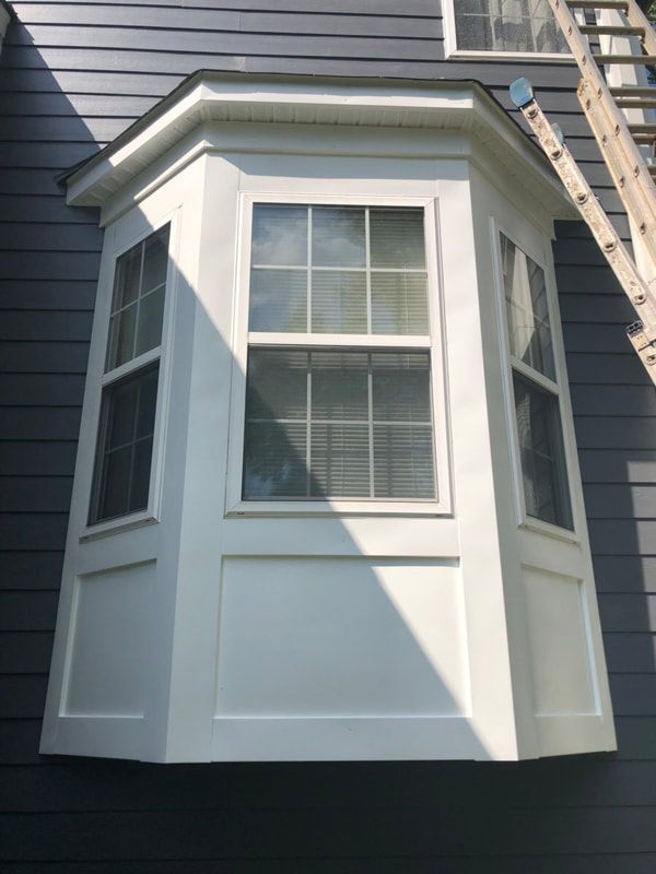 White bay window on a gray house, with a ladder leaning against the side.