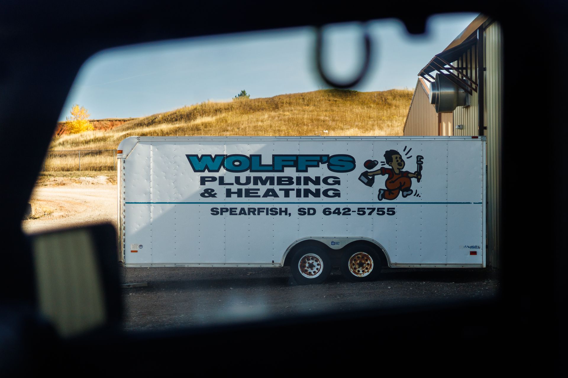 White Wolff's Plumbing & Heating trailer in Spearfish, SD. Seen through a vehicle window against a hillside.