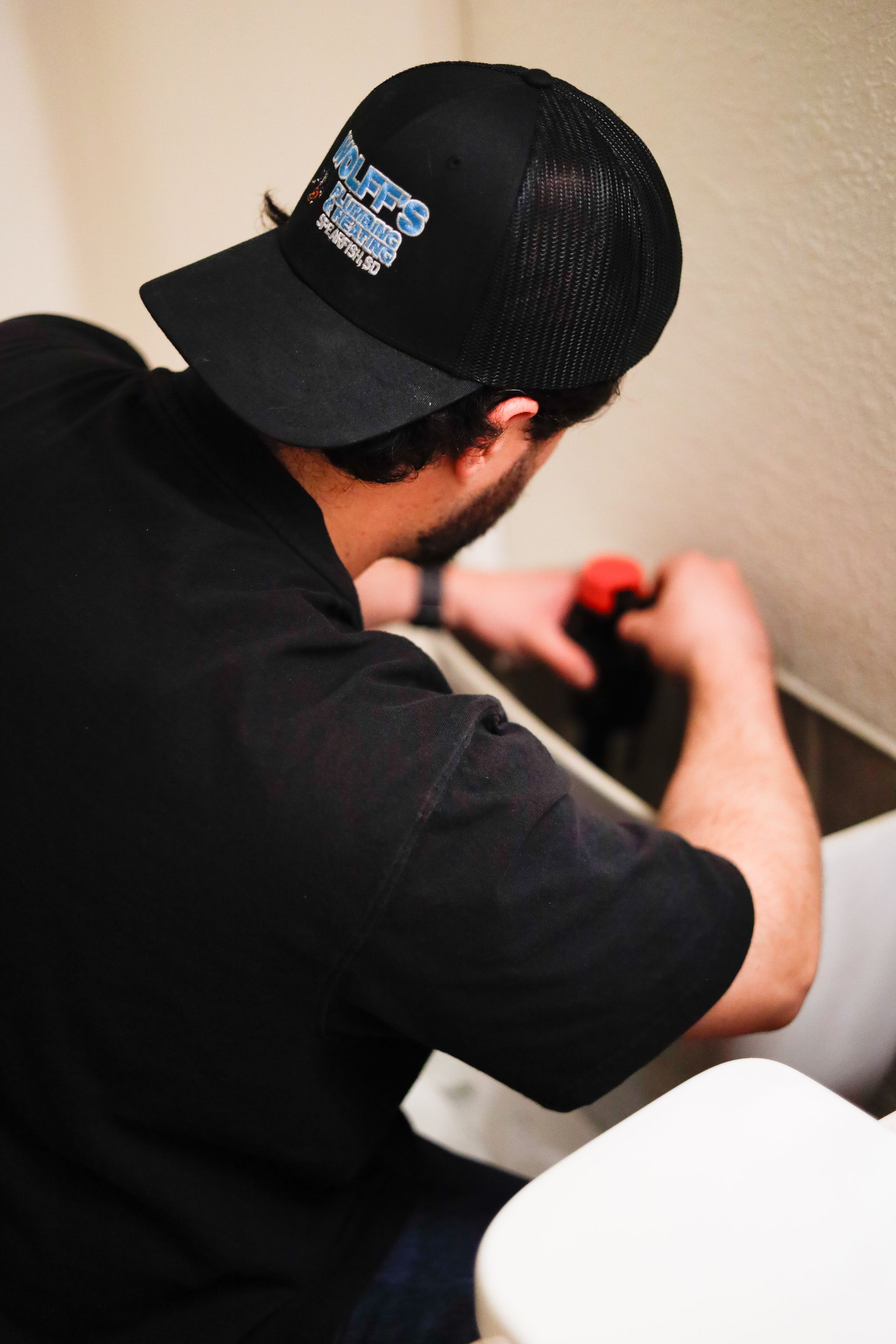 Man in black shirt and cap using a drill near a toilet.