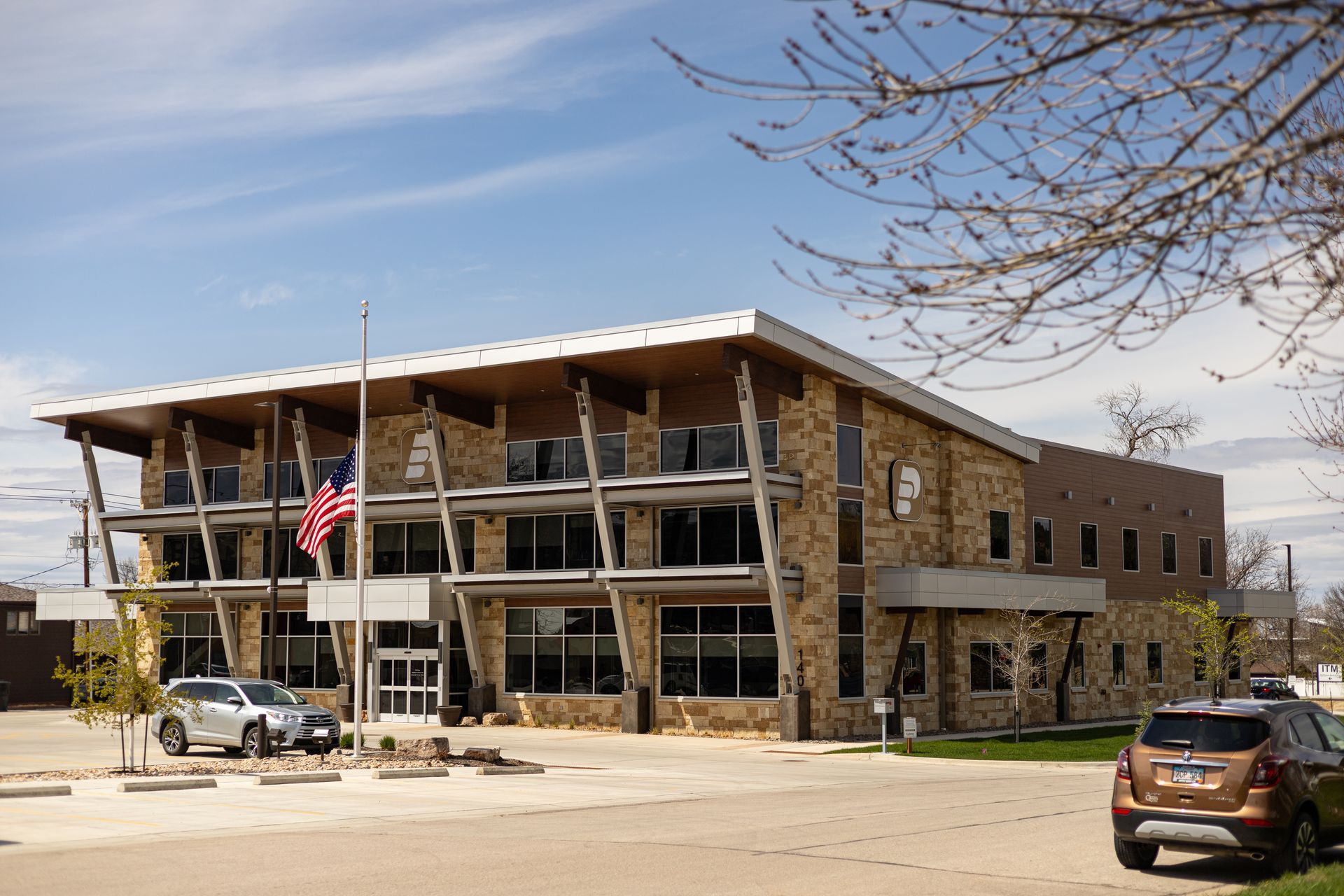 Two-story building with stone and glass facade, American flag, parked cars, clear sky.