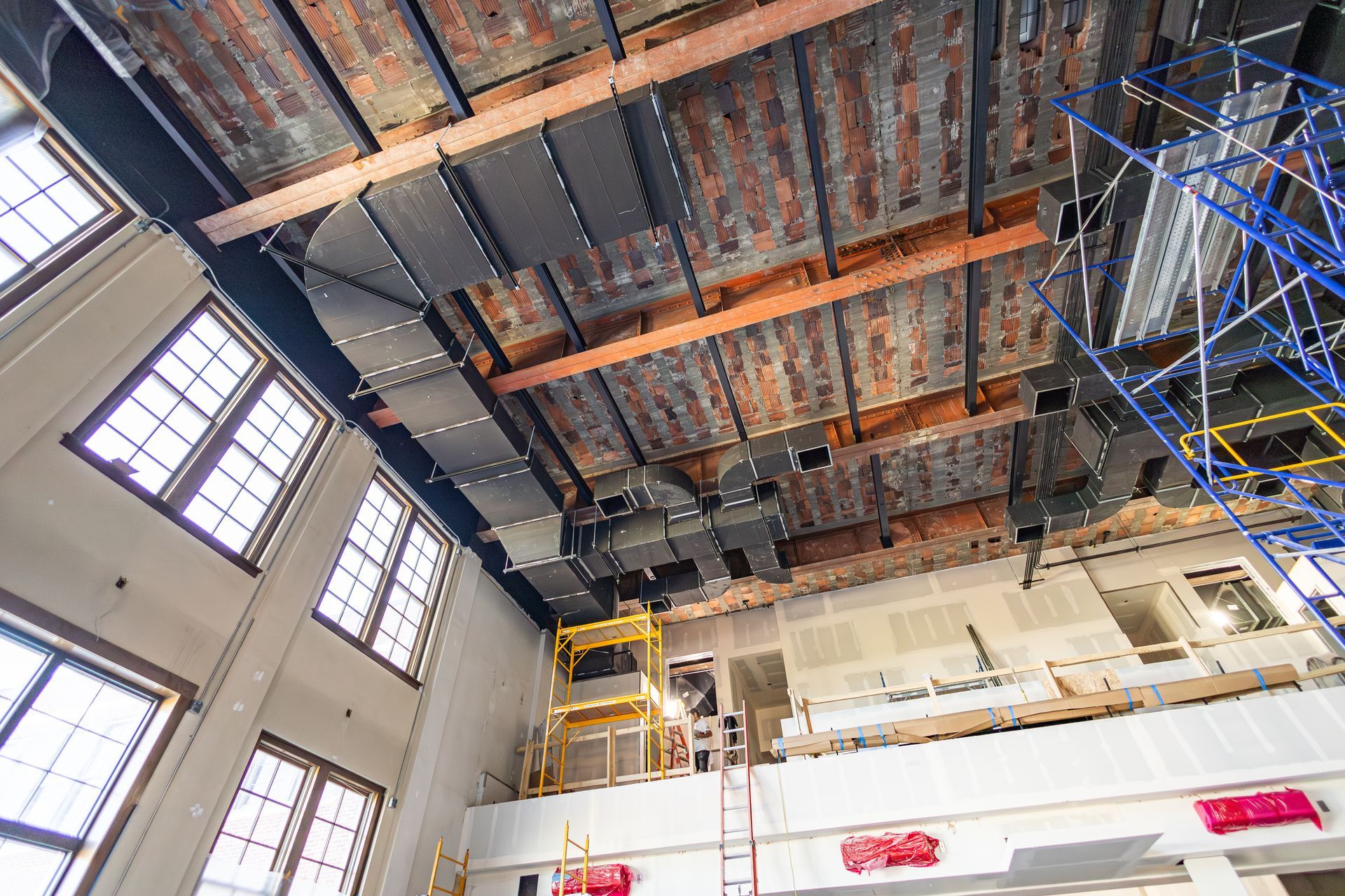 Construction site interior: exposed ceiling with ductwork, scaffolding, and windows.