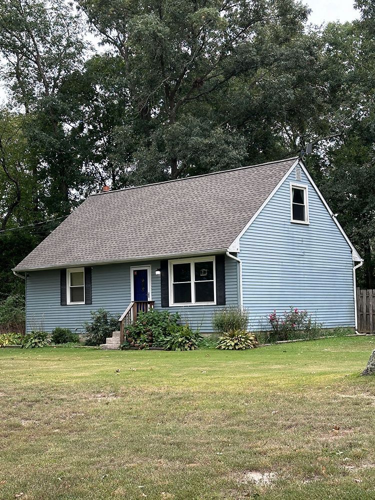 Blue-sided house with dark roof, front door, and two windows. Green lawn and trees in the background.