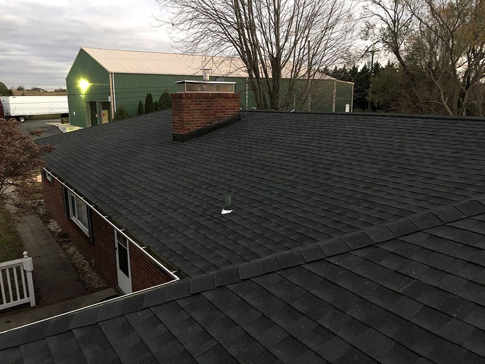 Black shingle roof with brick chimney, in front of a green warehouse, and white truck.