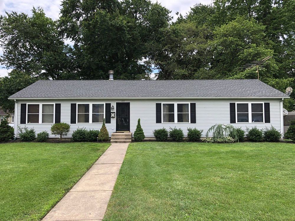 Ranch-style home with light siding, black shutters, and a grey roof, a pathway leads to the front door; surrounded by green lawn.