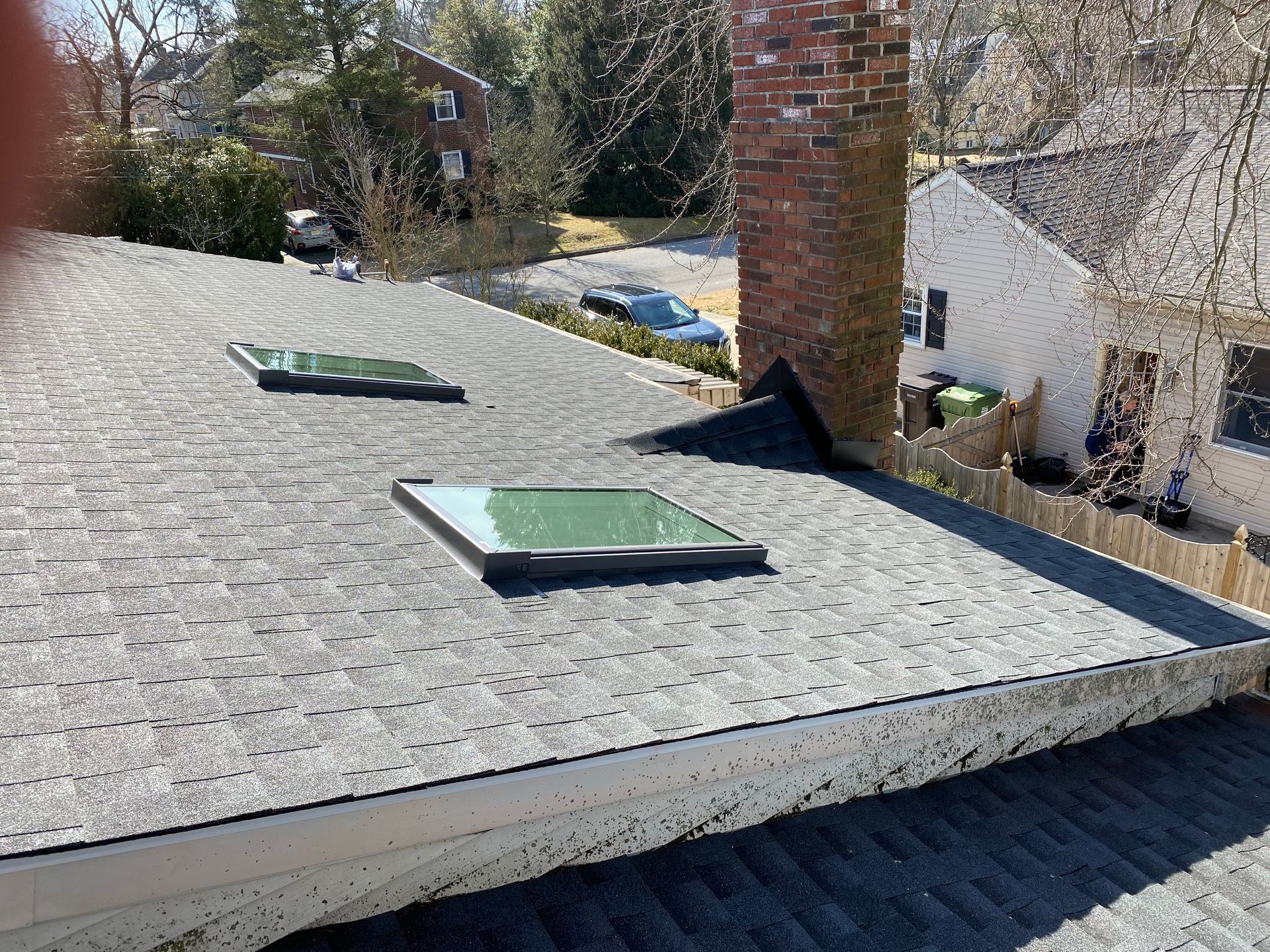 View of a shingled roof with two skylights and a brick chimney. Houses and trees in the background.
