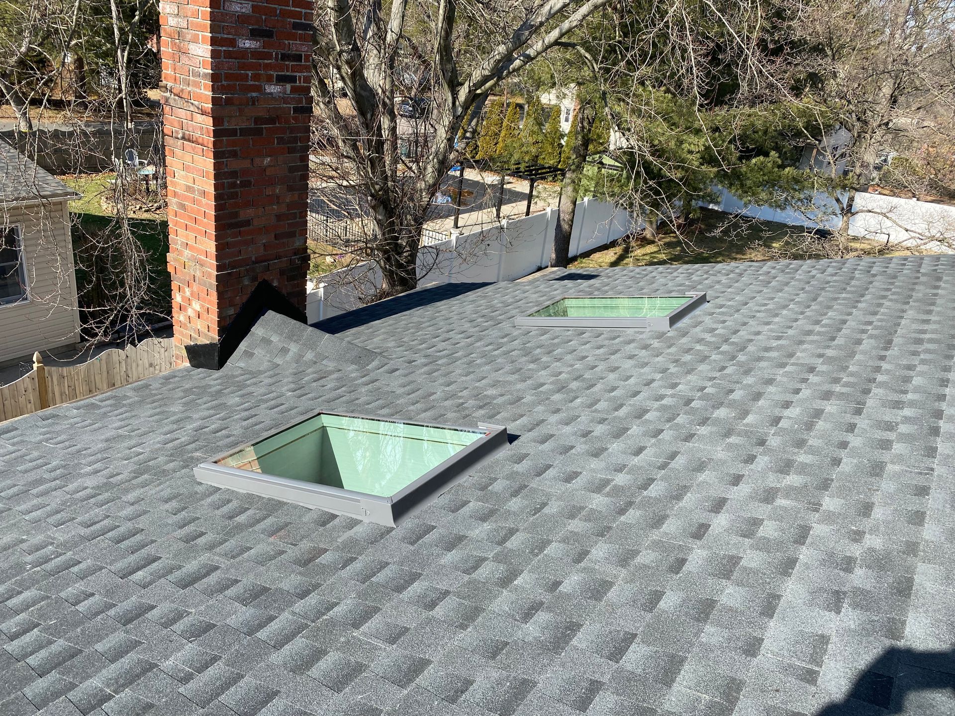 Gray shingle roof with two skylights, a brick chimney, and a fence in the background.