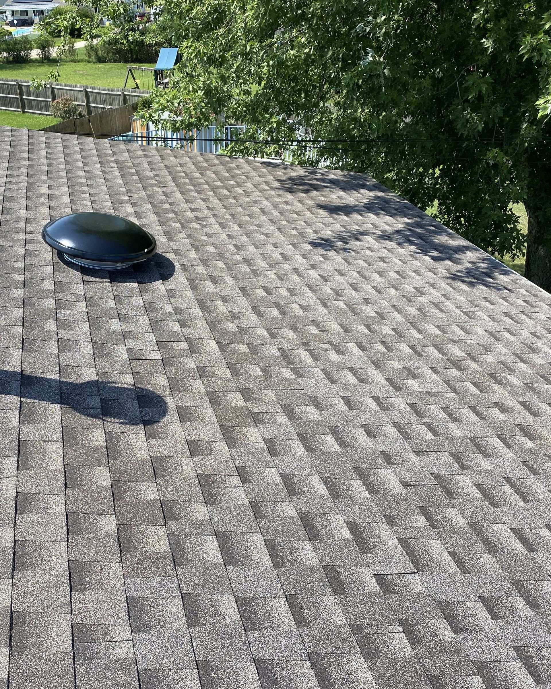 Overhead view of a shingled roof with a round, black skylight; trees and water are in the background.