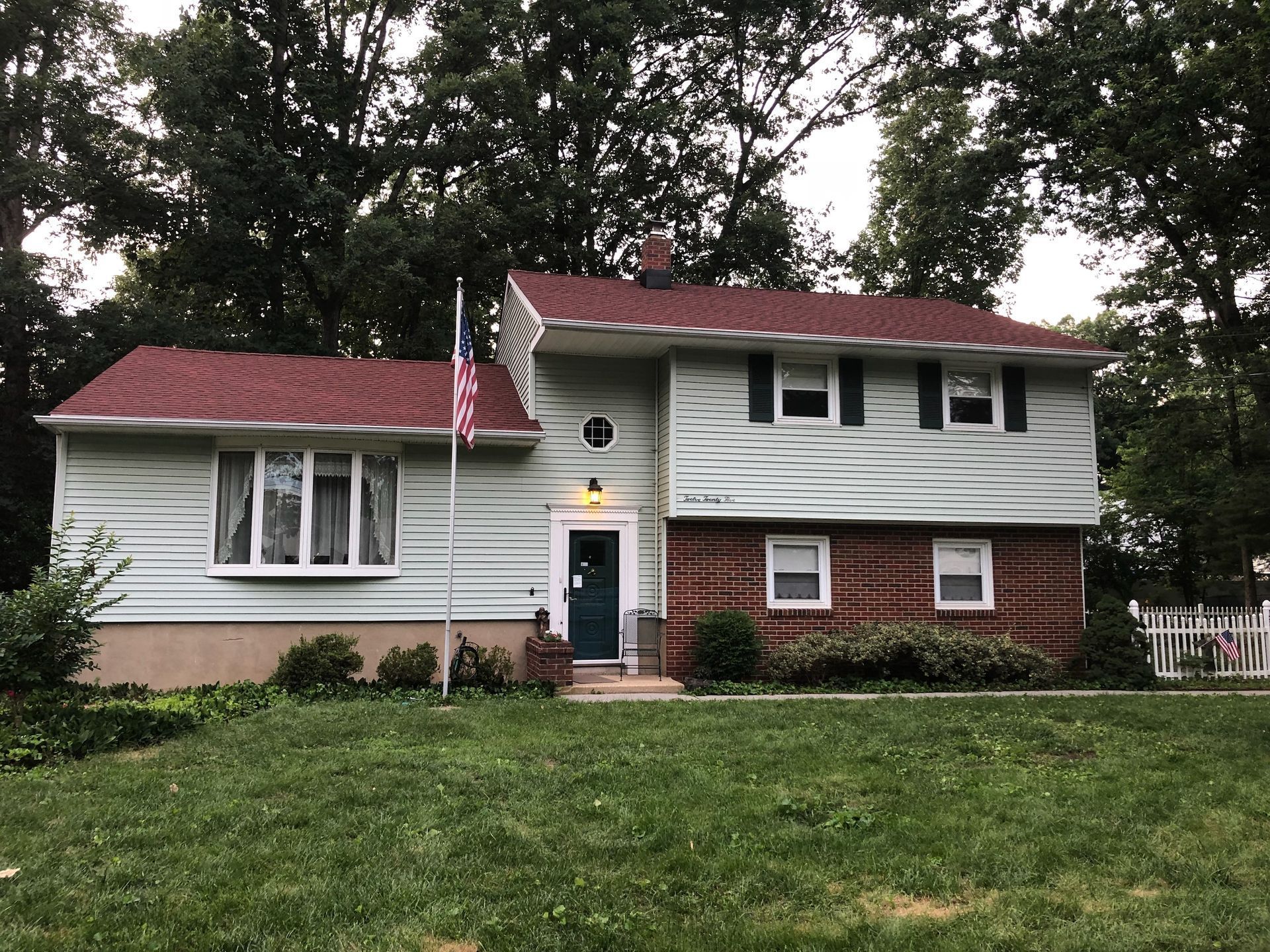 Two-story house with green siding, red roof, brick facade, and an American flag.