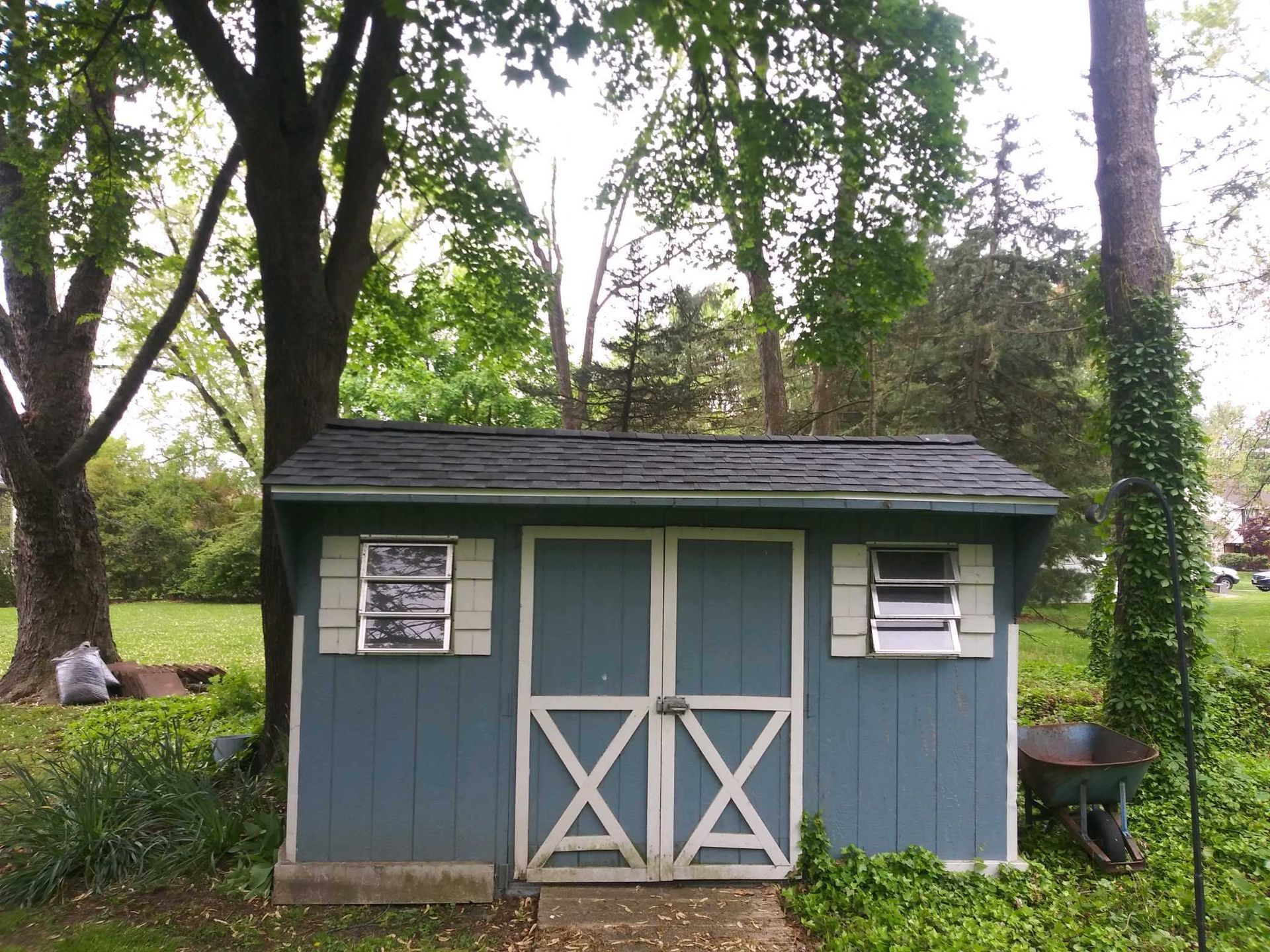 Blue shed with white trim and double doors in a yard with trees.