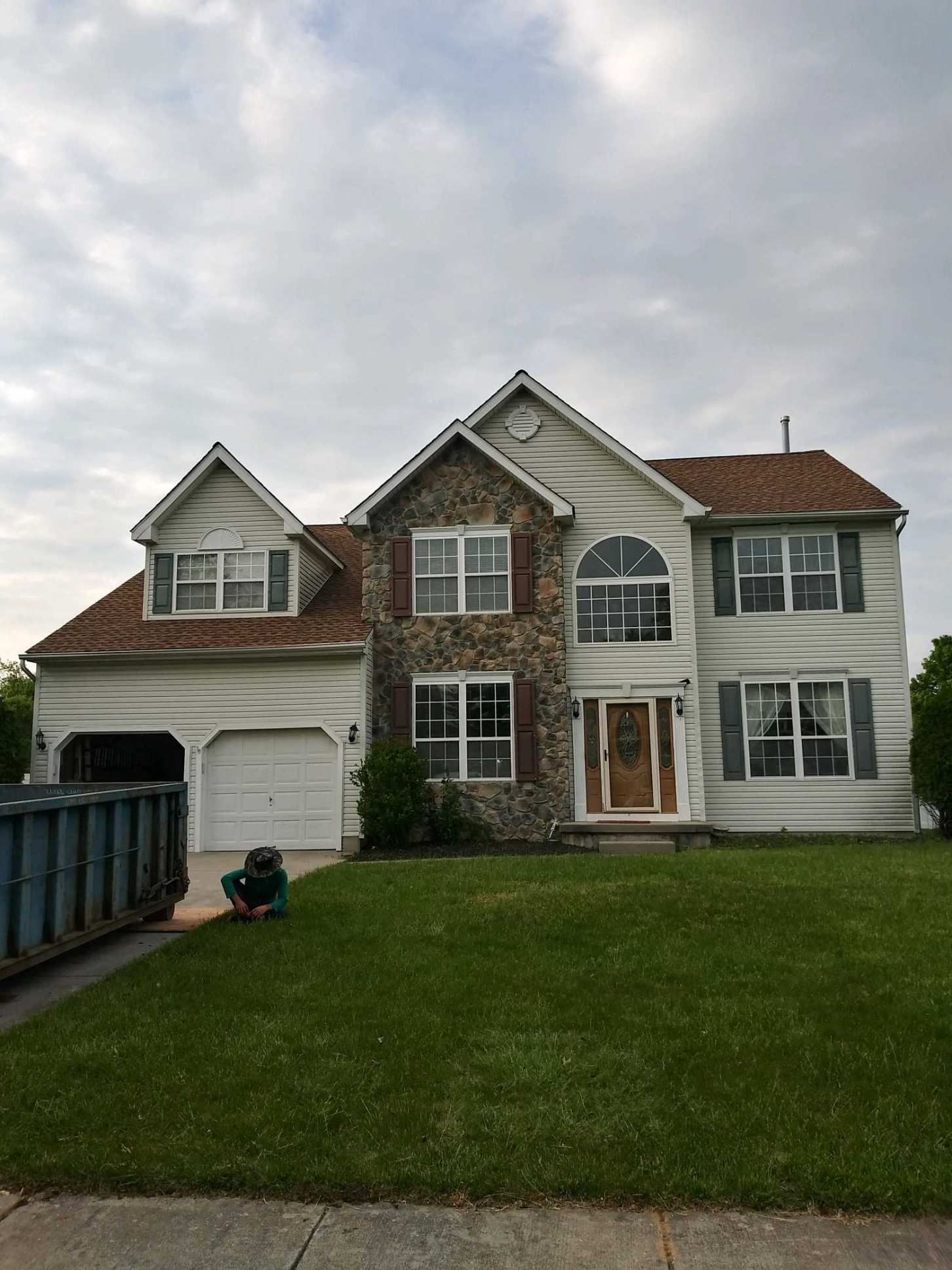 Two-story house with stone facade, brown shutters, and green lawn. A dumpster sits in the driveway.
